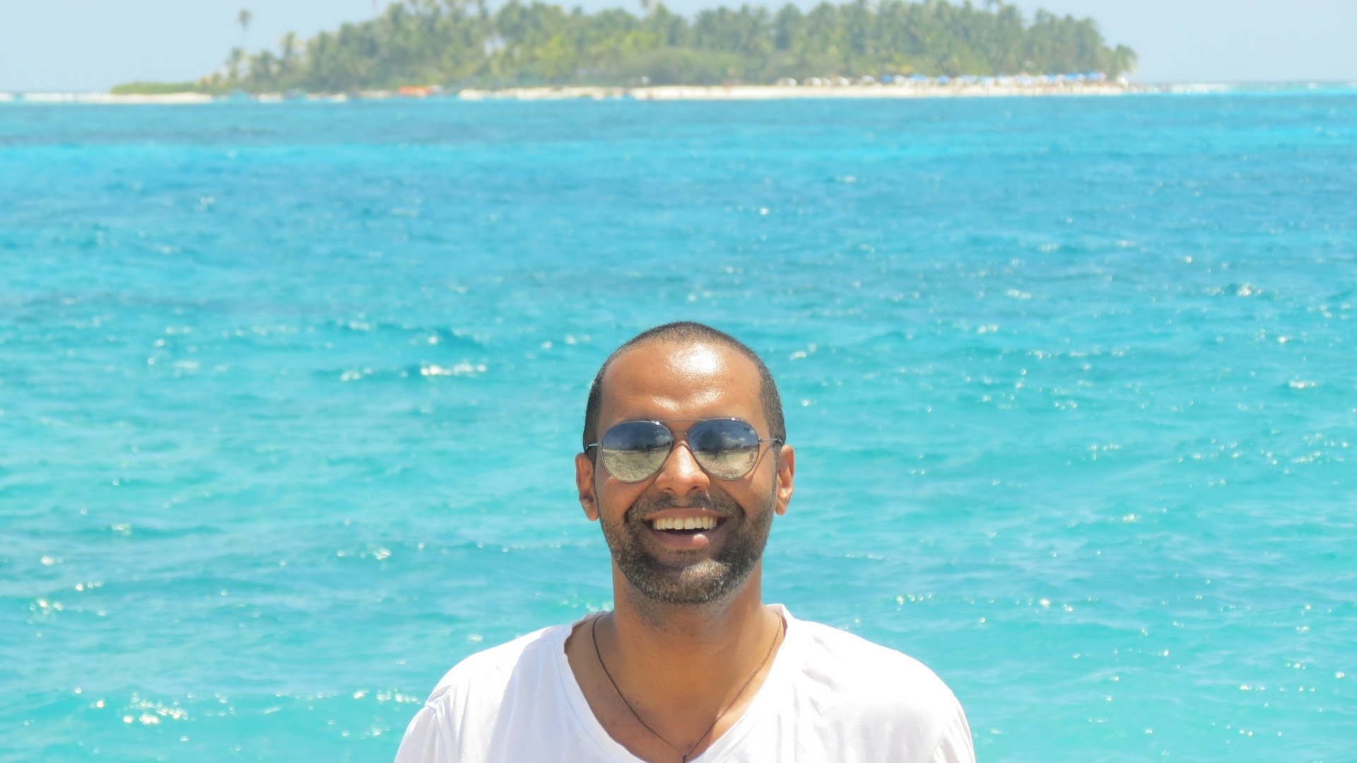 A man with wavy brown hair and a beard is standing in front of a shelf filled with various household items. He is wearing a light blue polo shirt and has a neutral expression.